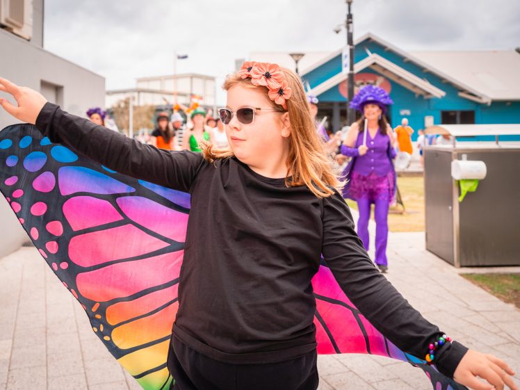 Image of A child wearing sunglasses, a flower headband, and colourful butterfly wings spreads their arms at an outdoor event. People in vibrant costumes and hats are visible in the background.