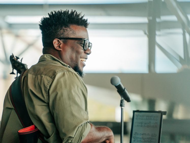Image of A smiling musician with glasses stands near a microphone, holding a guitar. He wears an olive green shirt and looks to the side. A music stand with sheet music is visible in the foreground.