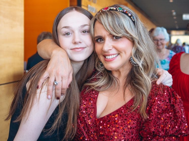 Image of Two women smiling and posing together indoors. The woman on the right wears a sparkly red dress and jewelled headband, while the younger woman on the left has straight brown hair and a black top. They appear happy and close.