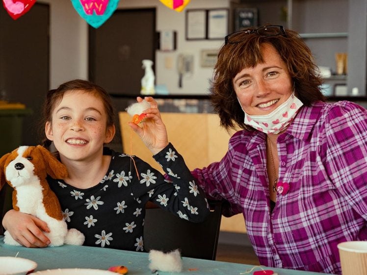 Image of A smiling woman and a young girl sit at a table doing crafts. The girl holds a stuffed dog and some stuffing, while the woman, wearing a checked shirt and lowered face mask, also smiles at the camera.