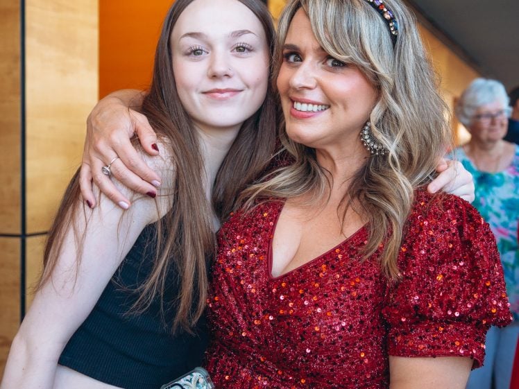 Image of Two women pose together, smiling. The woman on the left has long brown hair and wears a black sleeveless top, whilst the woman on the right has wavy blonde hair, a jewelled headband, and a sparkly red dress. Other people are blurred in the background.