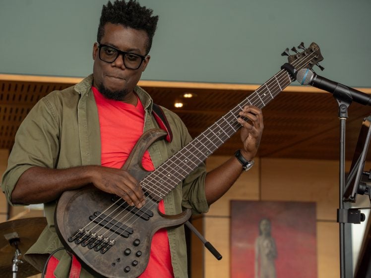 Image of A musician wearing glasses and a red shirt under an olive jacket plays a six-string electric bass guitar. A microphone and drum kit are nearby, with art on the wall in the background.