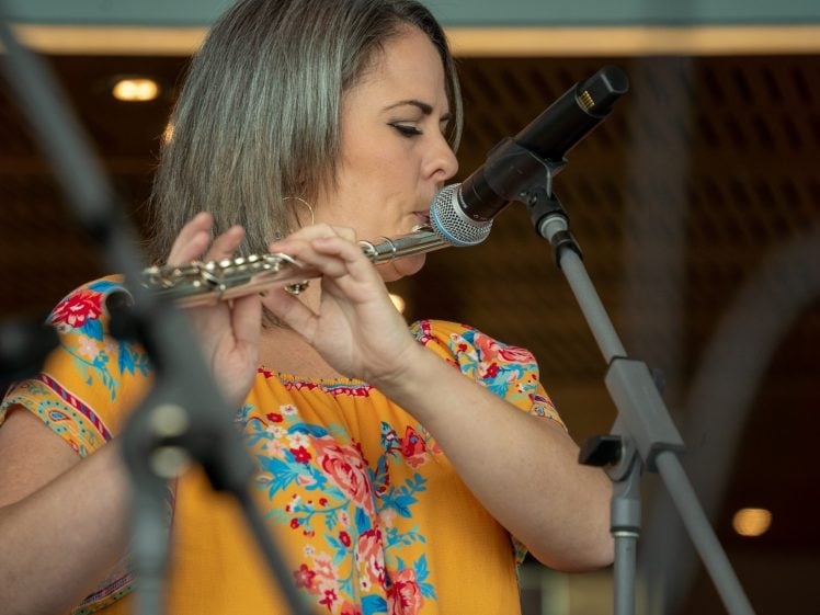 Image of A woman in a yellow floral blouse plays the flute and sings into a microphone on stage.
