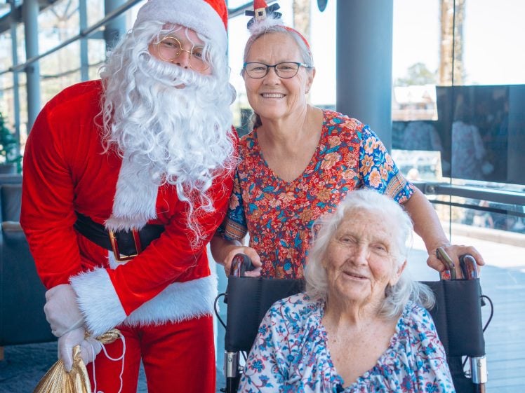 Image of A person dressed as Father Christmas stands beside two smiling elderly women, one in a wheelchair, inside a bright, sunlit building with large windows. The standing woman wears reindeer antlers and a floral top.