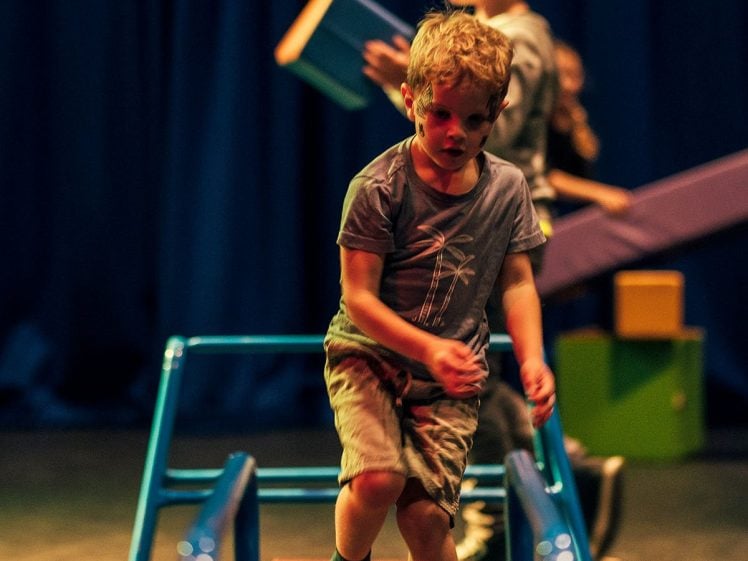Image of A young boy walks carefully on a wooden balance beam in a play area, whilst another child in the background holds a large foam block. The lighting is warm and the background is dark blue.