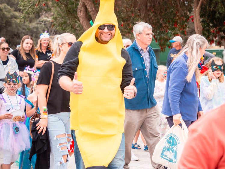 Image of A man dressed in a banana costume walks outdoors among a crowd, smiling and giving two thumbs up. People around him, including children in fancy dress, look cheerful. Trees and bright daylight set a festive atmosphere.