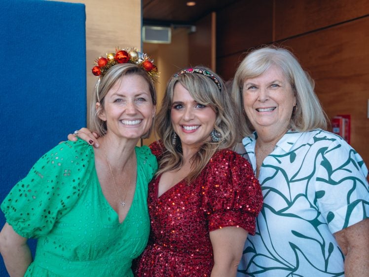 Image of Three women smiling together indoors. The woman on the left wears a green dress and a festive headband with red baubles, the middle woman wears a sparkly red dress, and the woman on the right wears a white dress with a green pattern.