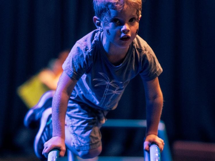 Image of A young child with face paint climbs over gym bars under blue stage lighting, looking focused. The background is dark and blurred.