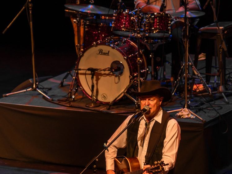 Image of A man in a cowboy hat sings and plays guitar on stage, with a drummer and another musician in the background, both wearing hats, illuminated by stage lights.