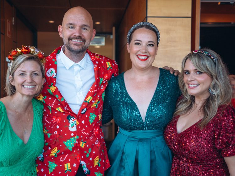 Image of Four smiling adults pose together at a festive event, dressed in holiday-themed attire with bright colours, sequins, and headbands, standing indoors in front of wood-panelled walls.