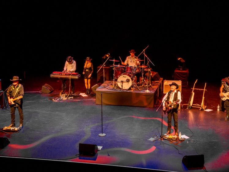 Image of A five-member band performs on a dimly lit stage. Musicians play guitar, keyboard, bass, and drums, and wear hats and stage outfits. Two microphones stand at the front, and instruments and equipment are visible.