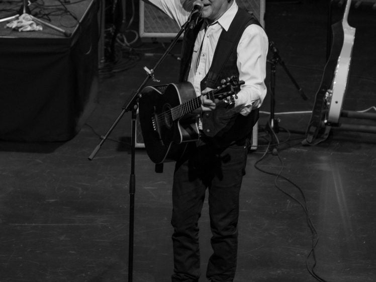 Image of A man in a cowboy hat plays an acoustic guitar and sings into a microphone on stage, raising one arm with fingers extended. Amplifiers, a guitar, and equipment are visible around him. The image is in black and white.