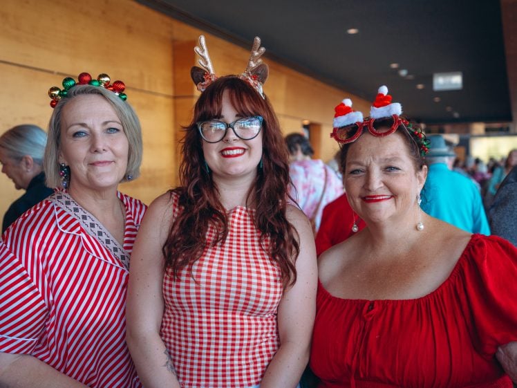 Image of Three women dressed in festive outfits and headbands, including Christmas decorations and reindeer antlers, smile together at an indoor holiday gathering.