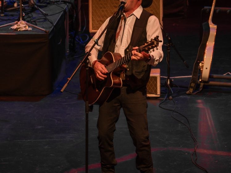 Image of A man in a cowboy hat sings and plays an acoustic guitar on stage. He stands near a microphone, with a pedalboard at his feet, a water bottle, and a sheet of paper on the floor. Musical equipment surrounds him.