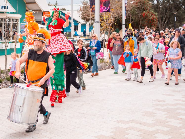 Image of People in colourful costumes, including a drummer in orange and performers on stilts, lead a lively parade. A cheerful crowd, some dressed up, follows along an outdoor path on a bright day.