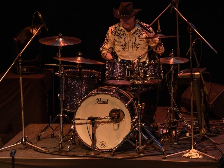 Image of A drummer wearing a cowboy hat and patterned shirt plays a drum kit on stage, surrounded by microphones and musical equipment, against a dark background.