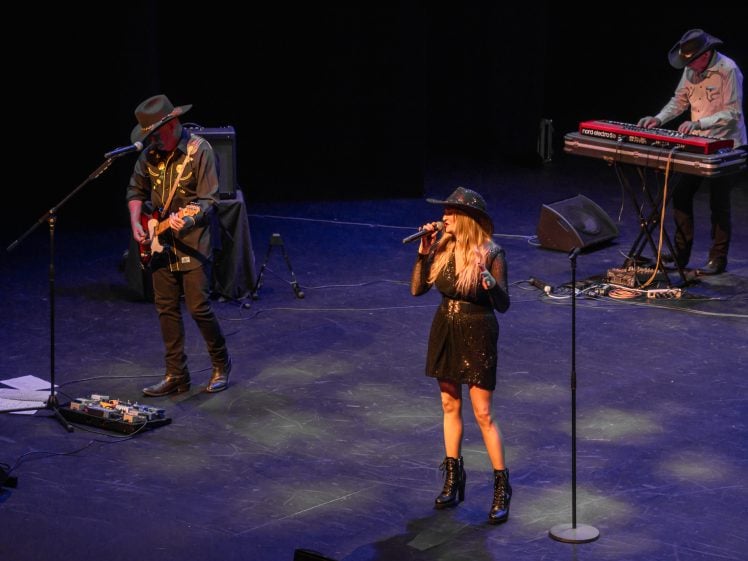 Image of Three musicians perform on stage: a woman sings at centre with a microphone, flanked by a guitarist on the left and a keyboard player on the right. All wear black outfits and cowboy hats. Stage lighting highlights them.