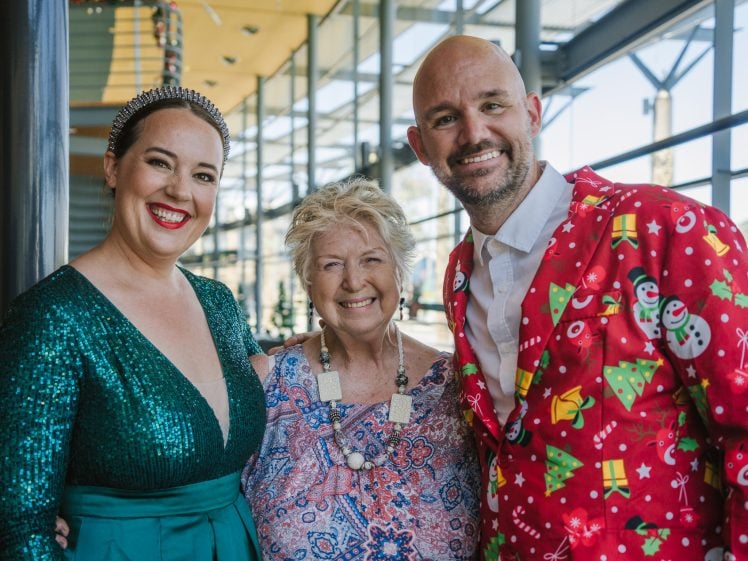 Image of Three smiling people pose together indoors. The woman on the left wears a sparkly green dress, the woman in the centre wears a patterned blouse, and the man on the right wears a festive Christmas suit with present and snowman designs.