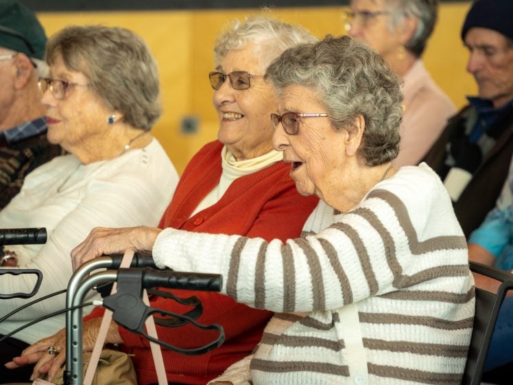 Image of A group of elderly people sit closely together, smiling and laughing. One woman in the foreground holds onto a zimmer frame, while others wear glasses and light jumpers. The mood appears cheerful and lively.