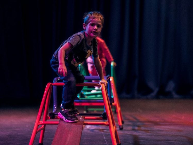 Image of A young boy in a grey shirt and trainers climbs over a red play structure on stage, with another child blurred in the background against a dark curtain.