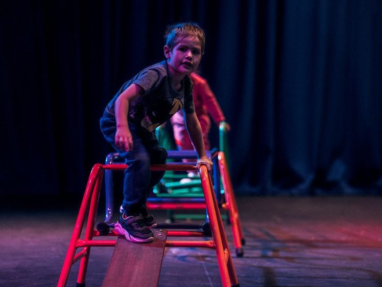 Image of A young boy balances on a red climbing frame on a stage with dark blue curtains, while another child waits behind him. The scene is dimly lit with colourful lighting.