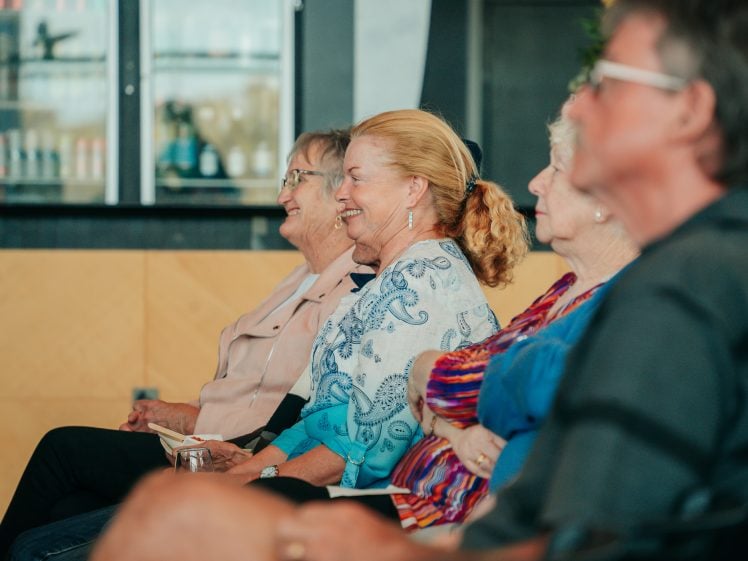 Image of A group of older adults sits side by side, smiling and attentively looking forward, as they enjoy an indoor event or presentation.