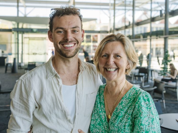 Image of A smiling man and woman stand together indoors in a bright, modern space with glass walls. The man wears a light shirt and the woman wears a green patterned dress. They pose closely, looking happy.