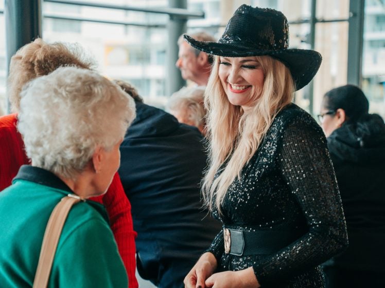Image of A smiling woman in a black sequin dress and cowboy hat talks with an older woman in a green jumper at a busy indoor event. Other people are gathered in the background.