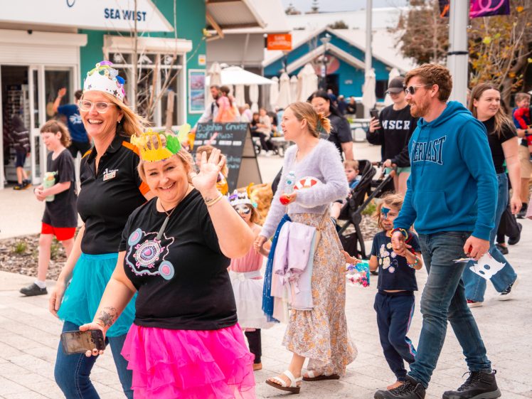 Image of A cheerful group of people, some wearing colourful paper crowns and tutus, walk together in a lively outdoor event. Others, including children, follow behind along a paved street near shops.