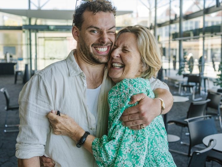 Image of A smiling man and woman stand indoors, hugging each other closely. The woman, wearing a green patterned dress, laughs joyfully while the man, in a light shirt, grins widely. Empty chairs and large windows are in the background.