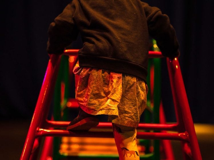 Image of A young child with bunches climbs up a red and green climbing frame ladder, wearing a dark jumper and a skirt with animal prints. The background is dark, creating a dramatic contrast with the lit child.