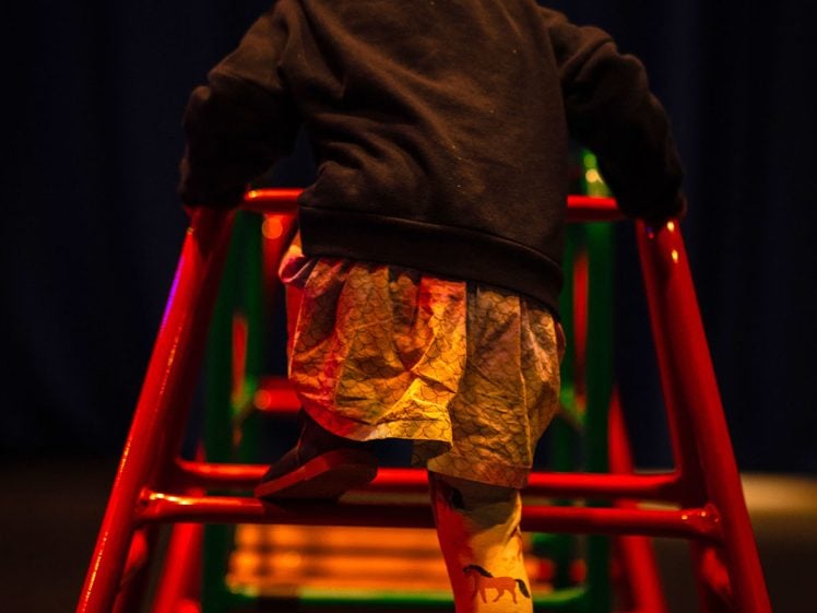 Image of A young girl with plaits climbs a red and green metal climbing frame, wearing a dark jumper, a skirt, and tights with animal prints. The background is dark, drawing focus to the child’s activity.