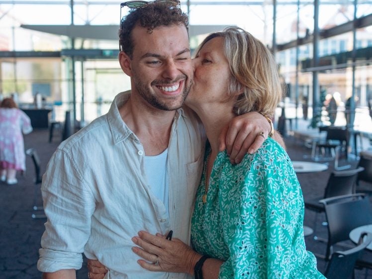 Image of A woman in a green patterned dress kisses a smiling man on the cheek whilst hugging him. They stand in a bright, modern indoor space with large windows and scattered chairs.