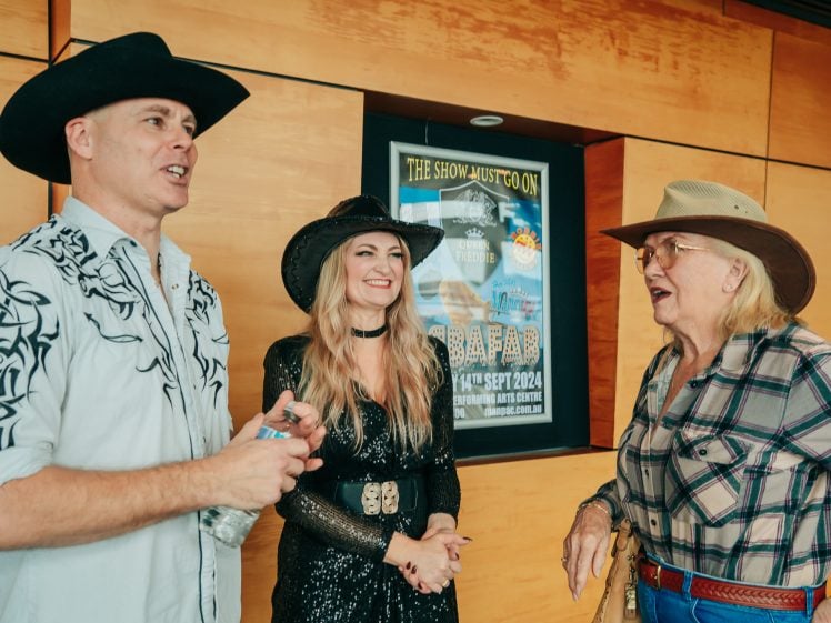 Image of Three people in cowboy hats and western outfits are smiling and talking indoors. A poster is visible behind them on a wooden wall. One person holds a water bottle while another wears glasses and a checked shirt.