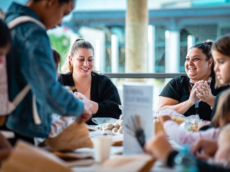 Image of A group of people, including adults and children, sit around a table enjoying a craft activity together. They are smiling and engaged, with art supplies and paper bags spread on the table.