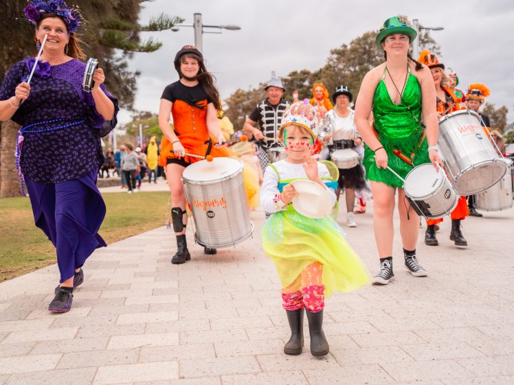 Image of A colourful parade features smiling adults and a child in bright costumes playing drums and dancing on a paved path, with trees and onlookers in the background. The child in front wears fairy wings and a crown.