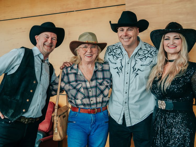 Image of Four adults dressed in Western-style clothing and cowboy hats stand together smiling in front of a light wood wall. Two men are on the left and middle right, and two women are on the middle left and right.