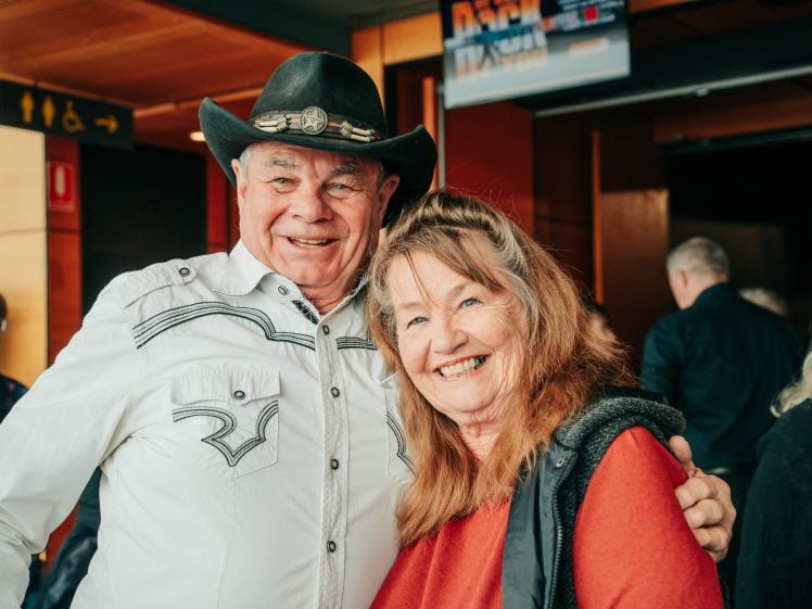 Image of An older man in a cowboy hat and white western shirt smiles with his arm round a woman in a red top. Both are happy and standing indoors with other people in the background.