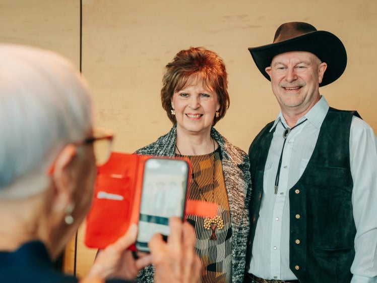Image of An older woman takes a photo on her mobile of a smiling man in a cowboy hat and waistcoat standing next to a woman with short brown hair, both posing against a beige background.