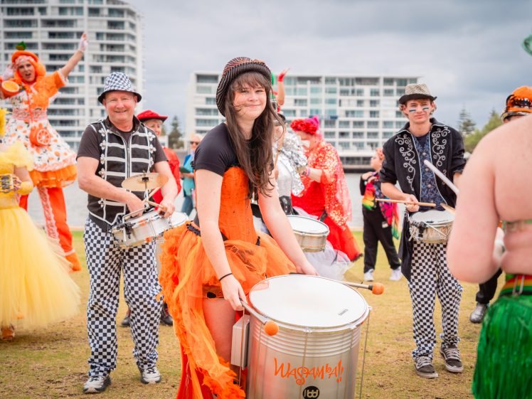 Image of A group of people in colourful, festive costumes play drums outdoors, with buildings in the background. The central figure wears an orange dress and smiles whilst playing, surrounded by others in bright and patterned outfits.