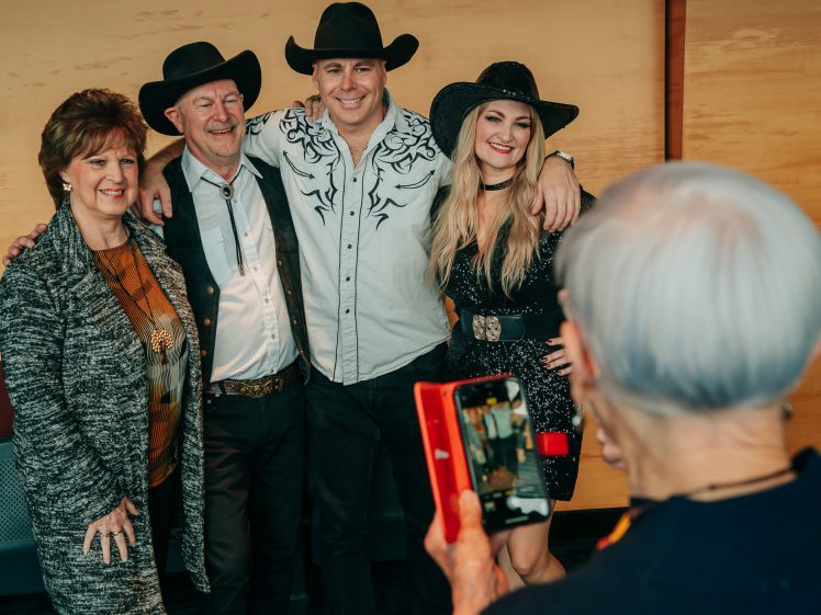 Image of Four people dressed in Western-style clothing pose and smile for a photo, while a person with short grey hair takes their picture using a red mobile phone. The background is a wooden wall.