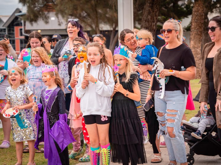 Image of A group of excited children and adults, many in colourful costumes and face paint, stand outdoors at an event. Some kids wear crowns and hold musical instruments, while adults watch and smile. Trees and buildings are visible in the background.