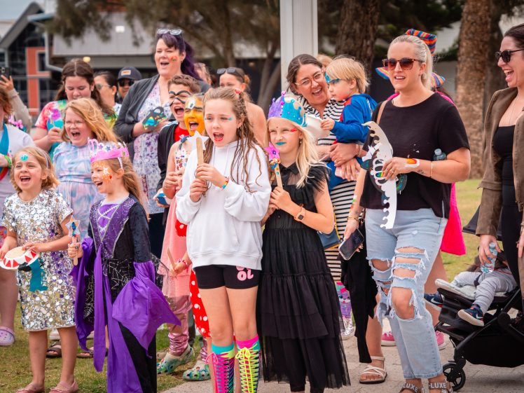 Image of A group of children and adults, some in colourful costumes and face paint, stand outside laughing and cheering at an event. A few children wear crowns and hold props, whilst adults watch and smile in the background.