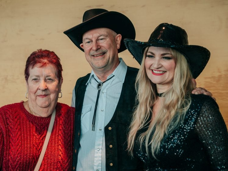 Image of Three people pose together, smiling. An older woman with short red hair wears a red jumper, while a man in a black cowboy hat and waistcoat stands beside her. A blonde woman in a black sequinned hat and dress stands on the other side.