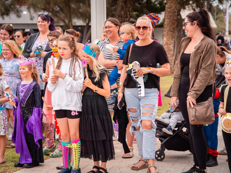 Image of A group of adults and children stand outdoors, many wearing colourful costumes, hats, and face paint, smiling and watching something off camera. Some children hold paper crafts, and a pushchair is visible among the group.