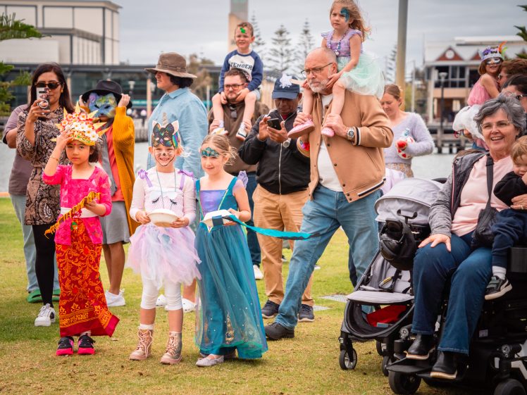 Image of A group of children in colourful costumes stand on grass, surrounded by adults, some taking photos. One child wears a red dress and crown, others are in fairy or princess outfits. People watch and smile; a toddler sits in a pushchair.