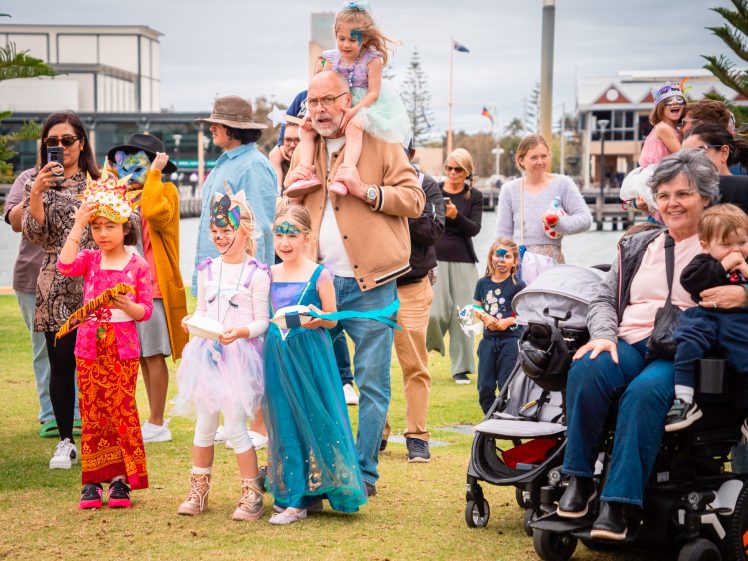 Image of Children in colourful costumes stand on grass with adults nearby, including a man carrying a girl on his shoulders and a woman in a motorised wheelchair holding a child. Several people are taking photos on a cloudy day.