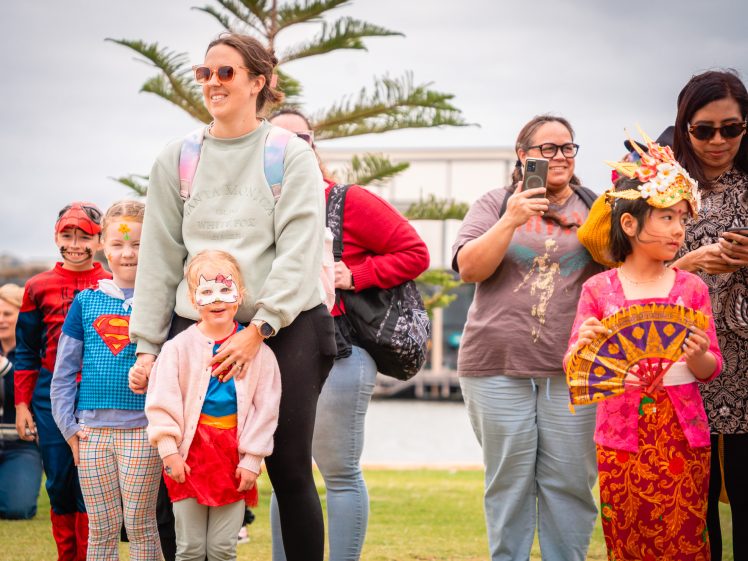 Image of A group of smiling children and adults, some in colourful costumes including superheroes and traditional attire, stand outdoors on grass near a tree on a cloudy day.