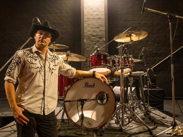 Image of A man in a cowboy hat and embroidered shirt stands beside a red drum kit on a stage, with microphones set up around the drums and a dark, textured wall in the background.