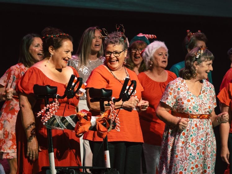 Image of A group of women, many wearing festive clothing and reindeer antlers, sing and smile on stage; two women in front stand by a decorated zimmer frame, all appearing joyful in a holiday-themed performance.